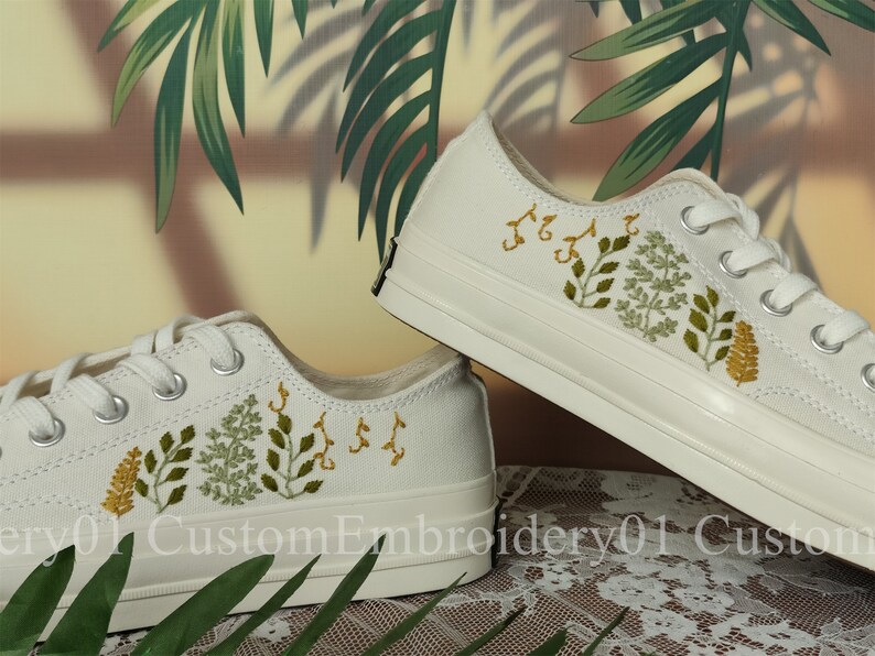 May include: A pair of white canvas sneakers with embroidered green and yellow floral designs. The shoes are a classic style with laces and a white rubber sole.