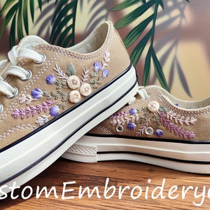 May include: A pair of beige canvas trainers with floral embroidery in shades of purple, pink, and green. The embroidery features delicate flowers and leaves.