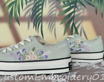 May include: A pair of light green canvas sneakers with white soles and black trim. The sneakers are decorated with embroidered flowers in shades of purple, pink, and white.