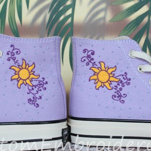 Peut inclure: Paire de baskets montantes lilas avec lacets et semelles blanches. Chaque chaussure présente un motif de soleil jaune brodé avec des vignes violettes. Les chaussures ont une surface texturée et une bande noire sur la semelle.