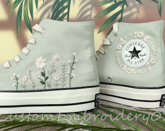 Könnte beinhalten: Ein Paar hellgrüne Converse-Sneaker mit hohem Schaft, die mit weißen gestickten Blumen und Blättern verziert sind. Das Converse-Logo ist an der Seite des rechten Schuhs sichtbar.