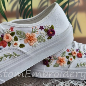 Puede incluir: Zapatillas blancas de lona con bordado floral en tonos rosa, naranja y morado. El bordado presenta flores, hojas y enredaderas. Las zapatillas tienen la palabra "VANS" impresa en el lateral.