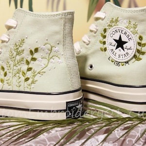May include: Pair of light green high-top sneakers with embroidered floral designs. The sneakers feature white laces, a black and white sole, and a Converse All Star patch. The floral embroidery includes green leaves and white flowers.
