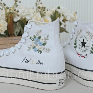 Może przedstawiać: Para białych, wysokich trampek Converse z indywidualnym haftem. Lewa trampka ma wz&oacute;r kwiatowy z niebieskimi i białymi kwiatami oraz napis "Lise & Ben". Prawa trampka ma zielony wz&oacute;r liści z czerwonym sercem i datą "01.19.25".