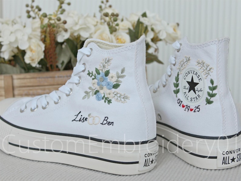Może przedstawiać: Para białych, wysokich trampek Converse z indywidualnym haftem. Lewa trampka ma wz&oacute;r kwiatowy z niebieskimi i białymi kwiatami oraz napis "Lise & Ben". Prawa trampka ma zielony wz&oacute;r liści z czerwonym sercem i datą "01.19.25".