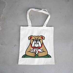 Puede incluir: Bolso tote de lona blanca con un diseño de bulldog de dibujos animados. El bulldog, en tonos marrones y rojos, sostiene naipes. El bolso tiene asas blancas y el bulldog está sentado sobre una superficie verde.