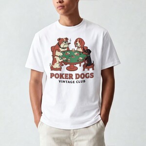 Puede incluir: Camiseta blanca con una ilustración de dibujos animados de perros jugando al póquer. La imagen muestra un bulldog, un beagle y un dachshund. El texto "POKER DOGS VINTAGE CLUB" está impreso en marrón.