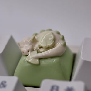 May include: A light green mechanical keyboard keycap featuring a sculpted, sleeping dragon figure in pale cream and soft green tones. The dragon has small wings and a row of rounded scales along its back, resting atop the smooth, square keycap surface.