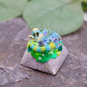 May include: A custom mechanical keyboard keycap featuring a sculpted, curled dragon in shades of blue, yellow, and green. The dragon rests on a textured green base atop a speckled, light gray, pyramid-shaped keycap base with a smooth finish.