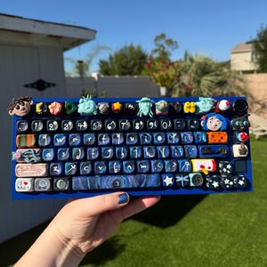 May include: A custom blue mechanical keyboard with unique keycaps. The keycaps feature various sculpted figures, patterns, and the text "I Mulch". The keyboard is held against a sunny outdoor backdrop.