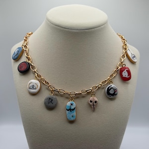 May include: A gold-tone chain necklace featuring several pendant charms. The charms are in various shapes and colours, including blue, red, and white, with painted designs. The necklace is displayed on a mannequin.
