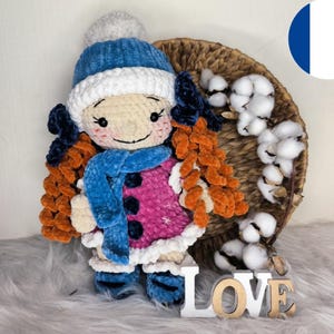 Peut inclure: Poupée au crochet faite à la main, vêtue d'un bonnet, d'une écharpe et de bottes bleus, et d'un manteau rose et blanc. La poupée a des cheveux roux et sourit. Le mot "LOVE" est épelé en lettres de bois en bas.