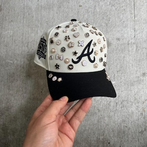 May include: A baseball cap with a white crown and a black brim. The crown is adorned with various decorative pins and the letter "A". The left side of the cap features a "150" patch. The cap is held by a hand.