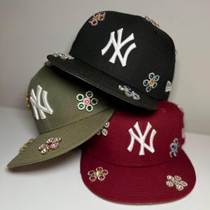 May include: Three baseball caps with the New York Yankees logo embroidered on the front. The caps are black, green, and red and are decorated with colorful jeweled flowers.