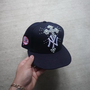 May include: A navy blue baseball cap with a white New York Yankees logo and a cross design embellished with white pearls.