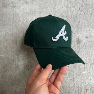Op de afbeelding: Een donkergroene baseballcap met een witte, getextureerde letter "A" op de voorkant. De cap heeft een gebogen klep en een gestructureerde kroon. Een klassiek accessoire voor casual wear.