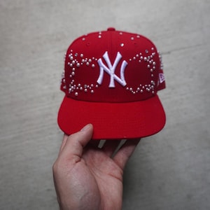 May include: A red baseball cap with a white New York Yankees logo and white rhinestones. The cap has a curved brim.
