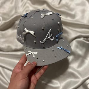 May include: A gray baseball cap with a white "A" embroidered on the front. The cap is decorated with white and blue bows and pearl-like embellishments.