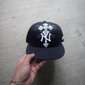 May include: A black baseball cap with a white New York Yankees logo and a cross design embellished with white pearls.