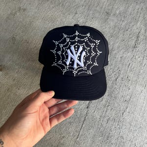 May include: A black baseball cap with a white spiderweb design and the letters "NY" in the center. The spiderweb and letters are covered in rhinestones.