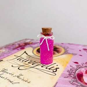 May include: A small clear glass bottle filled with vibrant pink glitter, sealed with a brown cork stopper and tied with a thin white string bow. The bottle rests on a purple book cover titled Fairyopolis A Flower Fairies Journal.