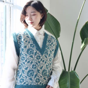 May include: A teal and cream-coloured knitted vest featuring a floral pattern. The vest has a V-neck and teal trim. The floral design is a repeating pattern of stylised flowers and leaves. The vest is worn over a white collared shirt.
