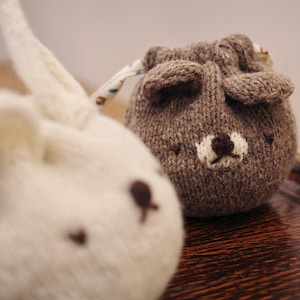 Puede incluir: Dos bolsas de punto con cordón, una blanca y otra marrón, cada una con forma de cabeza de animal. La bolsa blanca se asemeja a un conejo con detalles marrones, mientras que la bolsa marrón es un oso con hocico blanco y nariz marrón.