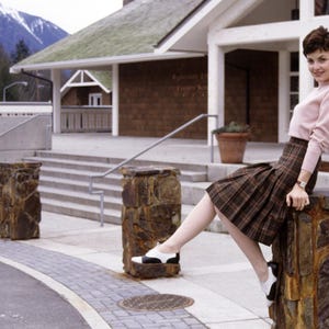 May include: A light pink long-sleeved sweater paired with a brown and black plaid pleated skirt. The outfit is completed with black and white saddle shoes. The clothing is worn by a person sitting on a stone pillar in front of a building.
