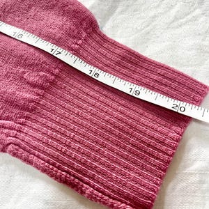 May include: A dusty rose knit sleeve with a ribbed cuff, positioned next to a white measuring tape that shows a length measurement extending from 16 to 20 inches along the fabric.