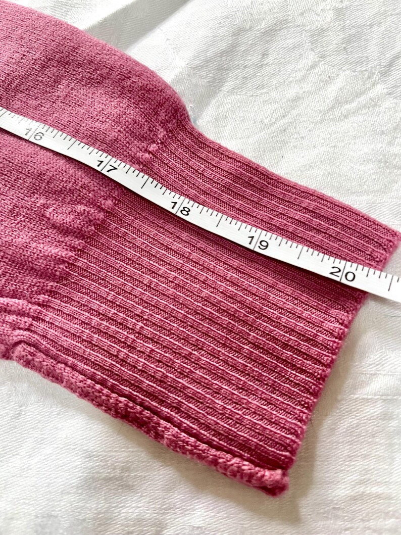May include: A dusty rose knit sleeve with a ribbed cuff, positioned next to a white measuring tape that shows a length measurement extending from 16 to 20 inches along the fabric.