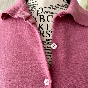 May include: A dusty rose knit polo shirt featuring a ribbed collar and a button-down front placket with two visible light pink buttons. The garment is displayed on a mannequin with black text printed on the fabric of the mannequin's neck area.