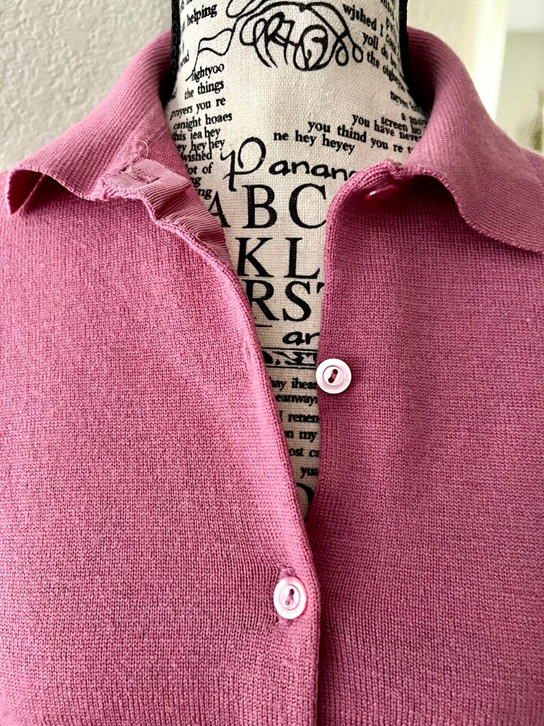 May include: A dusty rose knit polo shirt featuring a ribbed collar and a button-down front placket with two visible light pink buttons. The garment is displayed on a mannequin with black text printed on the fabric of the mannequin's neck area.