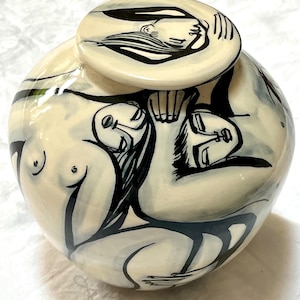 May include: A round ceramic jar with a matching lid, featuring a cream-colored glaze decorated with black line art illustrations of intertwined figures. The artwork depicts multiple faces and bodies in a continuous, flowing, and abstract style.