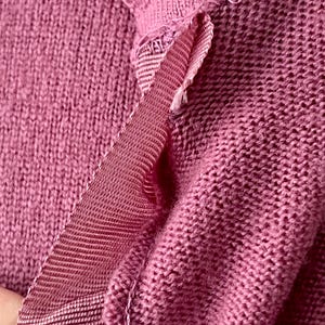 May include: A close-up view of a dusty rose knitted garment featuring a visible seam detail. The fabric has a textured, woven appearance with a matching grosgrain ribbon trim stitched along the edge, showing the construction of the knit material.