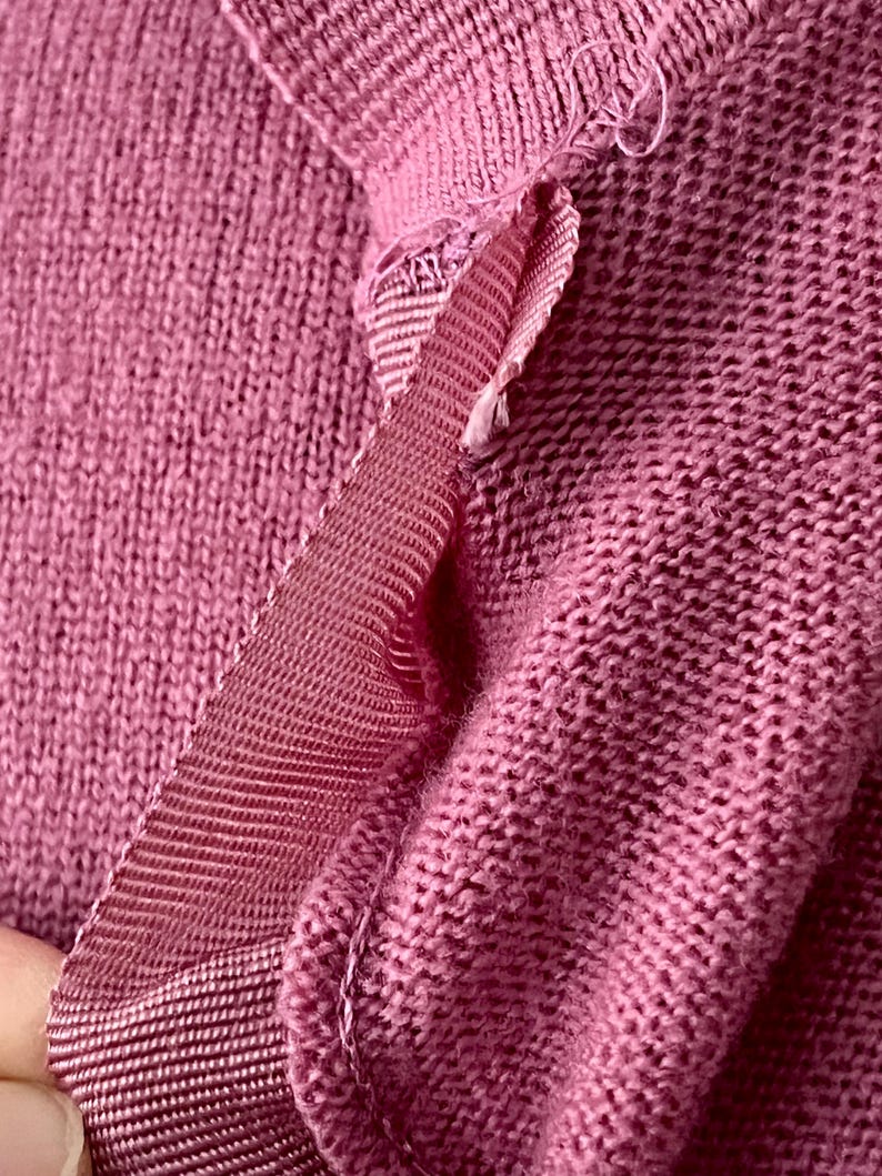 May include: A close-up view of a dusty rose knitted garment featuring a visible seam detail. The fabric has a textured, woven appearance with a matching grosgrain ribbon trim stitched along the edge, showing the construction of the knit material.