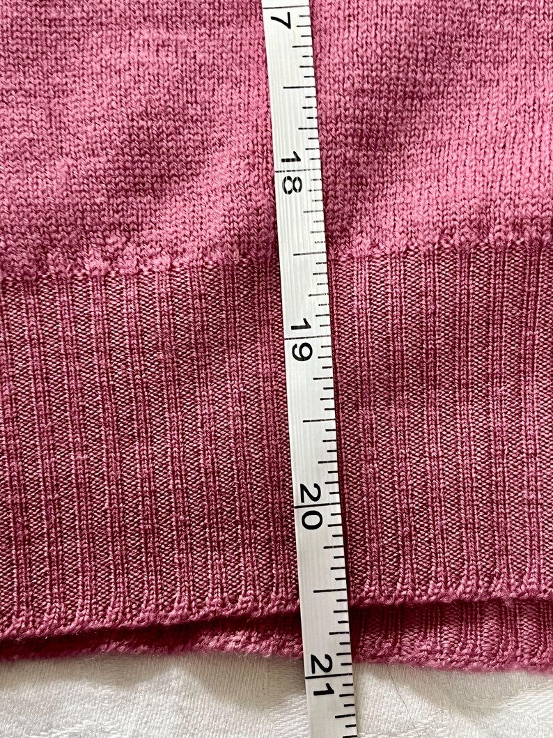 May include: A mauve-colored knit sweater featuring a ribbed hemline. A white measuring tape is placed vertically against the fabric, showing a length measurement extending from approximately 17 inches to 21 inches along the ribbed section.