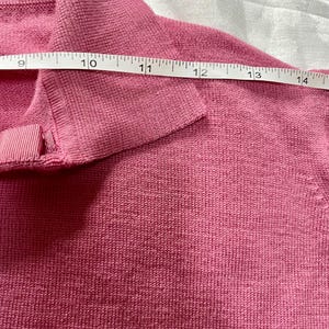 May include: A pink knit sweater with a folded collar and a small ribbon detail at the neckline. A white measuring tape is placed across the upper chest area, showing a measurement spanning from 9 to 14 inches across the garment's width.