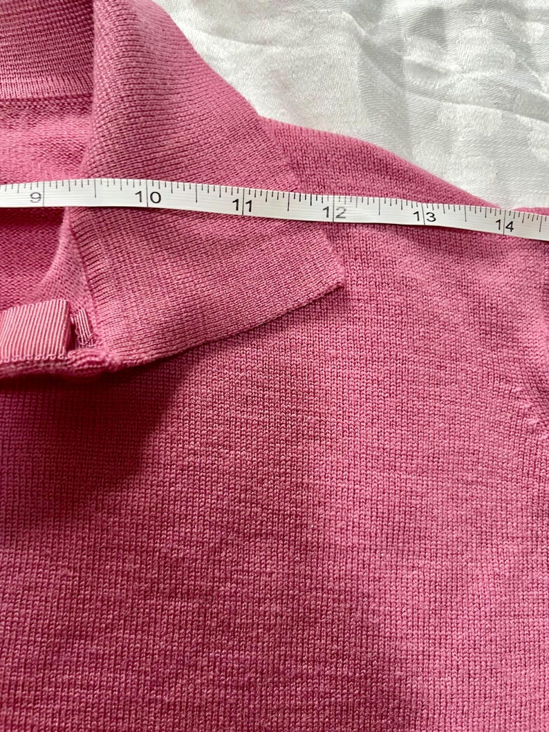 May include: A pink knit sweater with a folded collar and a small ribbon detail at the neckline. A white measuring tape is placed across the upper chest area, showing a measurement spanning from 9 to 14 inches across the garment's width.