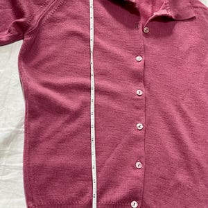 May include: A mauve button-down cardigan with a collar and long sleeves. A white measuring tape is placed vertically along the front, showing a length of approximately 21 inches. The garment features a row of light-colored buttons and a label.