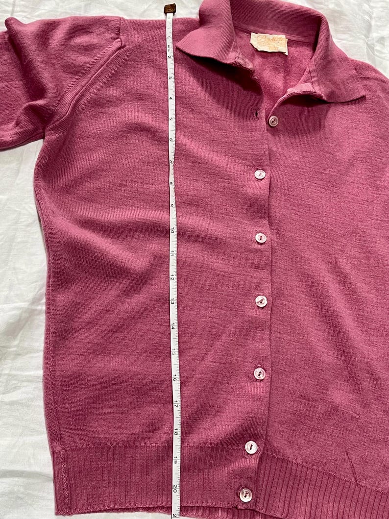 May include: A mauve button-down cardigan with a collar and long sleeves. A white measuring tape is placed vertically along the front, showing a length of approximately 21 inches. The garment features a row of light-colored buttons and a label.