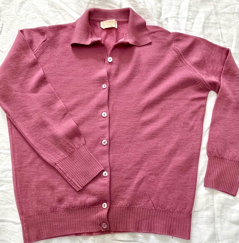 May include: A long-sleeved, mauve-colored knit cardigan featuring a collared neckline, a front button-down closure with six small white buttons, and ribbed cuffs and hem. The garment is made of a fine-gauge knit fabric with a consistent texture.