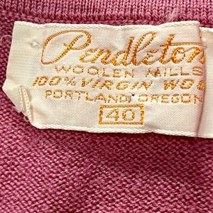 May include: A pink knit garment featuring a white fabric label with gold embroidered text that reads Pendleton Woolen Mills, 100% Virgin Wool, Portland, Oregon, and the number 40 enclosed in a rectangular box, stitched onto the textured fabric.