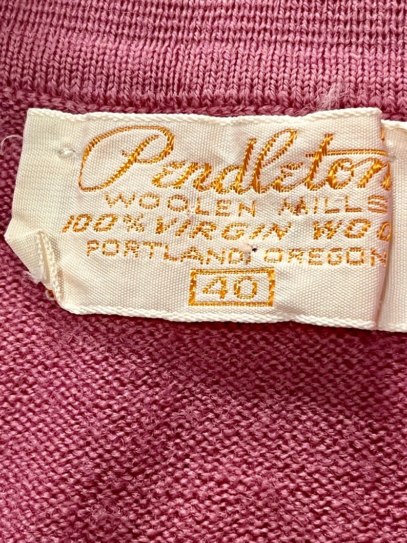 May include: A pink knit garment featuring a white fabric label with gold embroidered text that reads Pendleton Woolen Mills, 100% Virgin Wool, Portland, Oregon, and the number 40 enclosed in a rectangular box, stitched onto the textured fabric.
