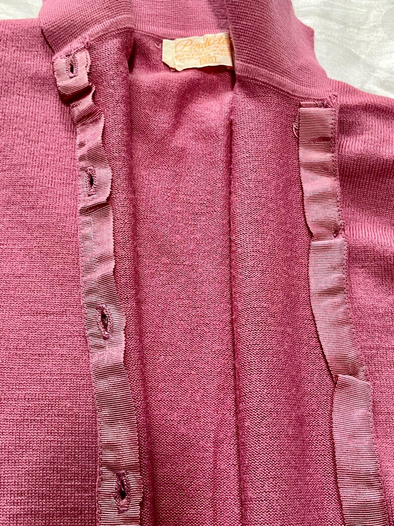 May include: A dusty rose knit cardigan featuring a button-down front with a matching grosgrain ribbon placket. The garment has a high collar and a Pendleton brand label at the inner neckline, displaying the text Pendleton and the number 40.