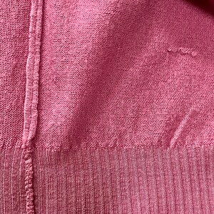 May include: A close-up view of a dark pink knit sweater featuring a vertical seam running down the left side and a ribbed hem at the bottom. The fabric has a consistent textured knit pattern with a small area of visible wear on the right side.