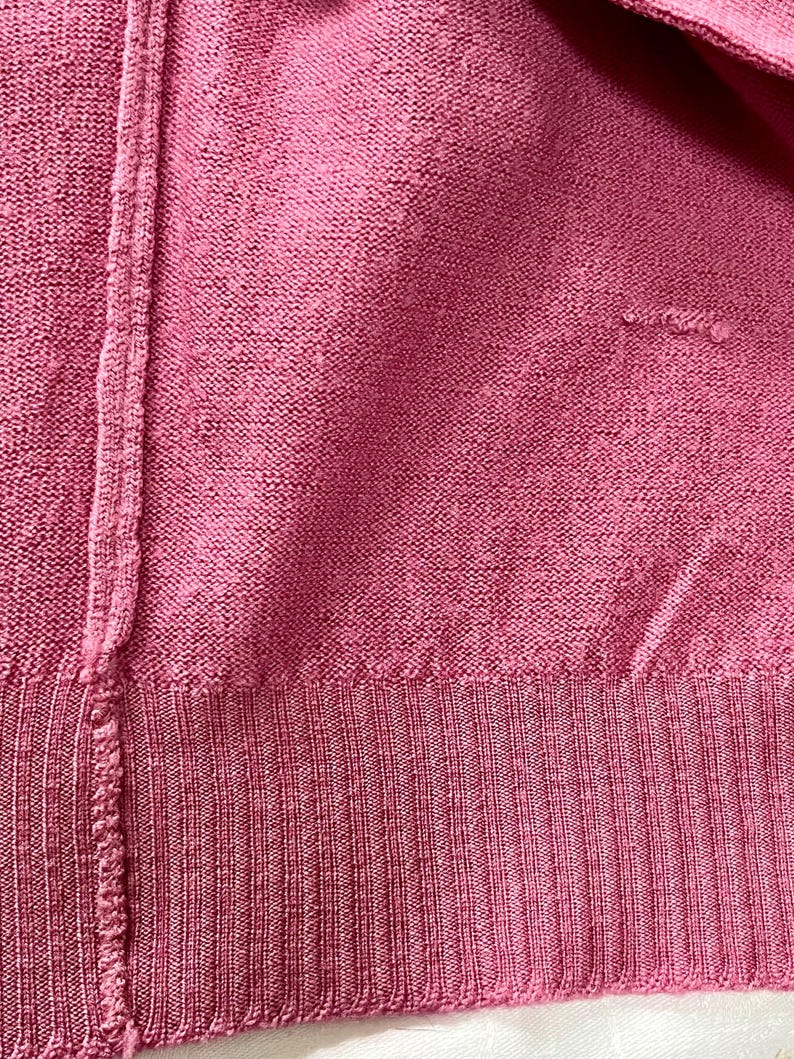 May include: A close-up view of a dark pink knit sweater featuring a vertical seam running down the left side and a ribbed hem at the bottom. The fabric has a consistent textured knit pattern with a small area of visible wear on the right side.