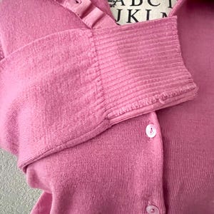 May include: A solid pink long-sleeved cardigan featuring a collared neckline, a button-down front with small white buttons, and ribbed cuffs. The garment is displayed on a mannequin with text printed on the neck area, including the letters ABC.