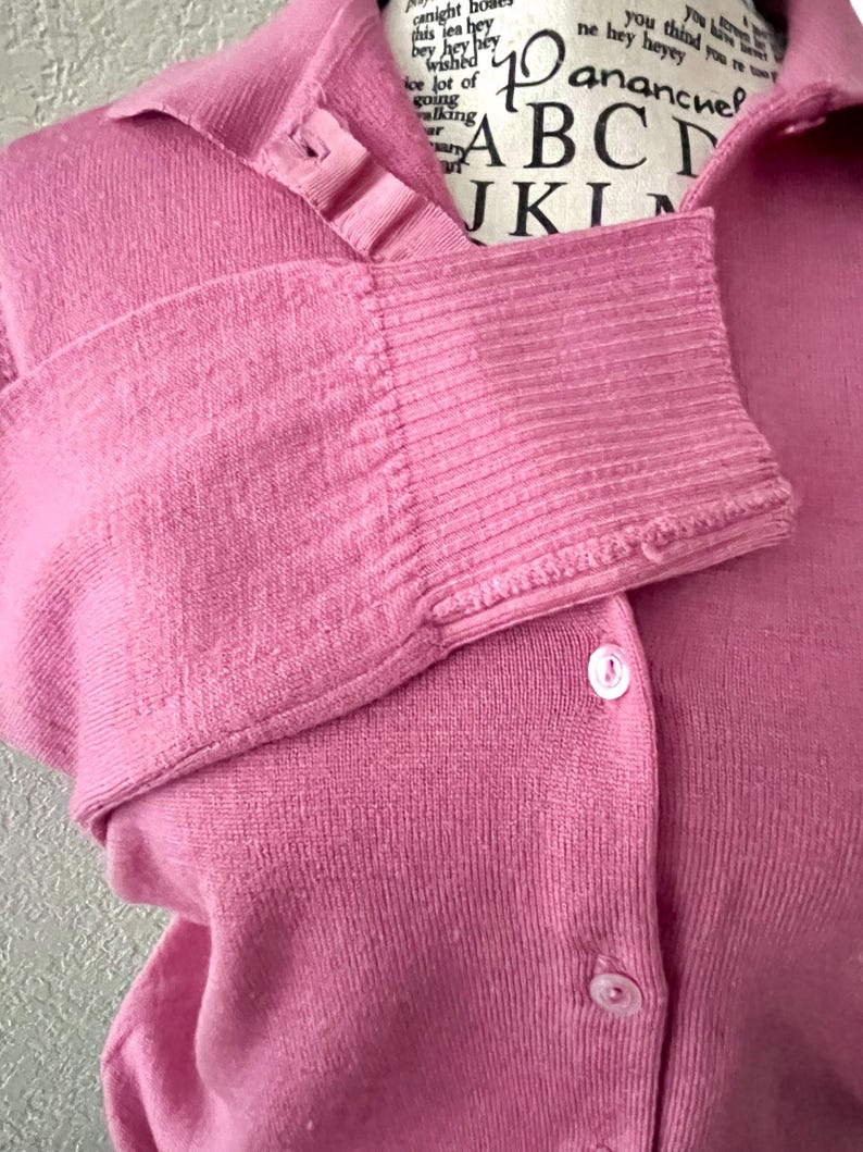 May include: A solid pink long-sleeved cardigan featuring a collared neckline, a button-down front with small white buttons, and ribbed cuffs. The garment is displayed on a mannequin with text printed on the neck area, including the letters ABC.