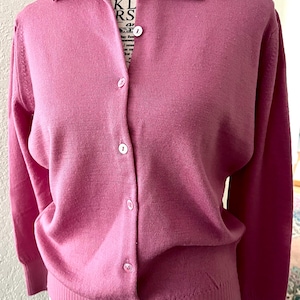 May include: A long-sleeved, button-down cardigan in a solid mauve-pink color. The garment features a classic collar, a vertical row of small white buttons down the center, and ribbed knit detailing along the cuffs and the bottom hemline.