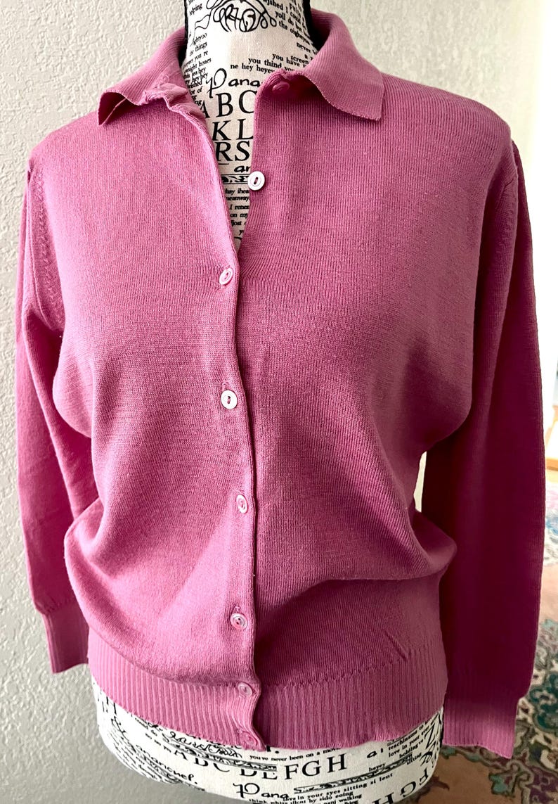 May include: A long-sleeved, button-down cardigan in a solid mauve-pink color. The garment features a classic collar, a vertical row of small white buttons down the center, and ribbed knit detailing along the cuffs and the bottom hemline.